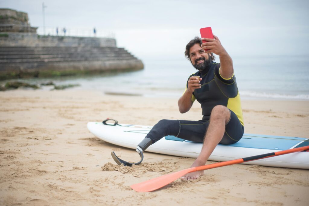 Man with a prosthetic leg sitting on a surfboard and taking a selfie
